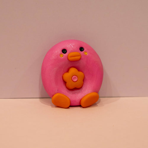 A small pink bird-shaped accessory with orange feet, beak, and flower detail, photographed against a plain light background.