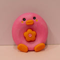 A pink, donut-shaped clay figure with an orange beak, orange feet, and a flower on its chest, set against a plain beige background.