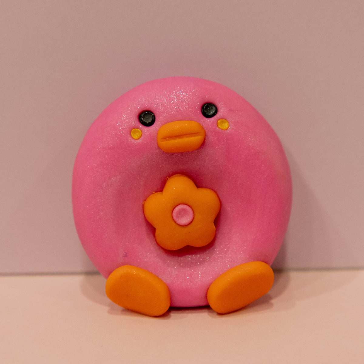 A pink, donut-shaped clay figure with an orange beak, orange feet, and a flower on its chest, set against a plain beige background.