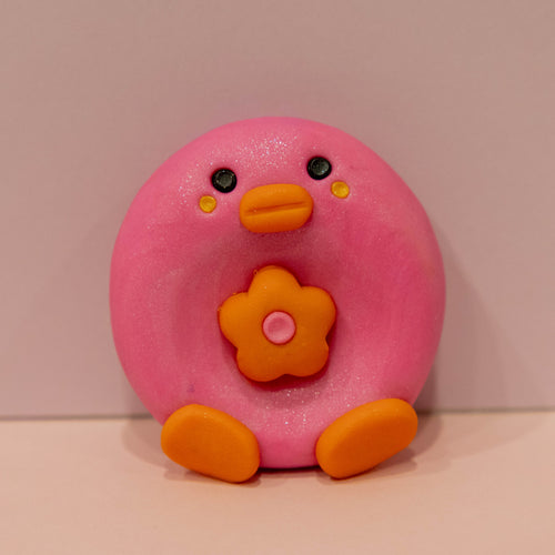 A pink, donut-shaped clay figure with an orange beak, orange feet, and a flower on its chest, set against a plain beige background.
