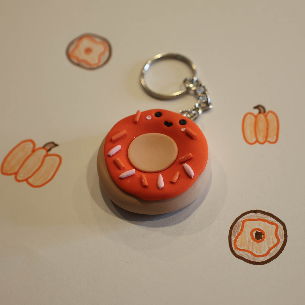A novelty keychain shaped like a donut with a smiling face, orange frosting, and white sprinkles, attached to a silver metal ring. The keychain is displayed on a white sheet of paper with small pumpkin and donut drawings scattered around it.