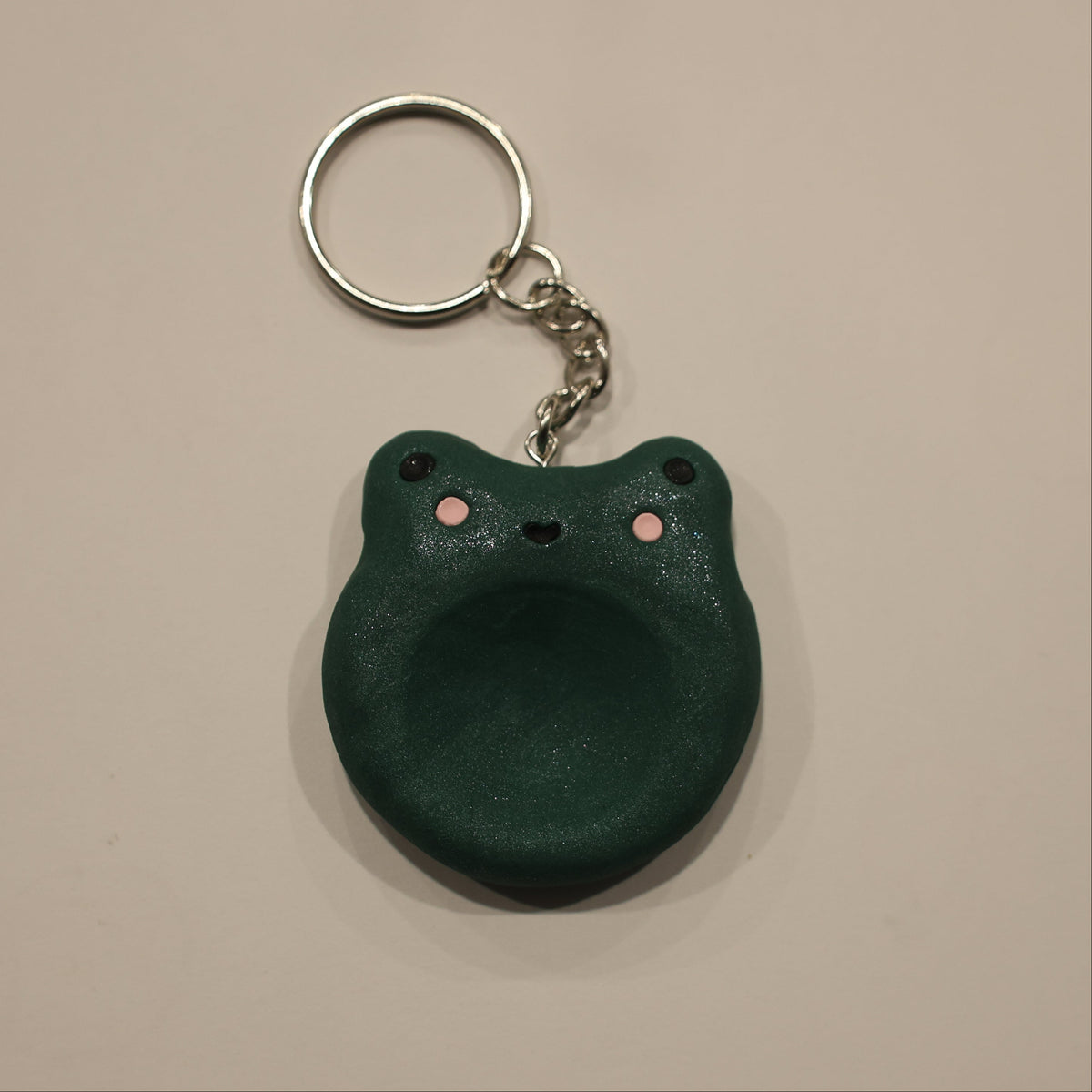 A dark green frog-shaped keychain with a silver metal split ring and chain, photographed on a plain beige background.